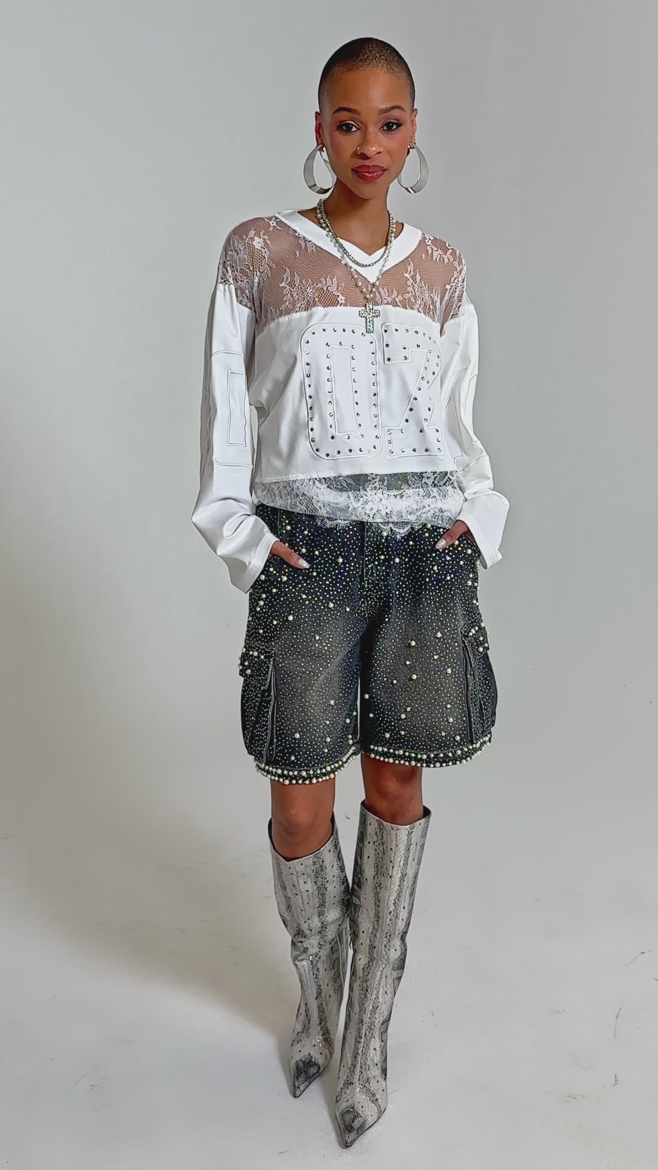 Video of woman modeling the WHO IS GETTING LUCKY TONIGHT JERSEY—a white long sleeve top with lace details—over a sheer lace shirt, paired with embellished pants. She stands against a plain background.