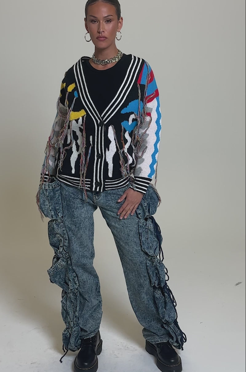 Video of woman modeling the SWEATER WEATHER CARDIGAN, a colorful oversized fringe cardigan, paired with a black top, chunky necklace, hoop earrings, and baggy blue jeans with ruffled sides against a light gray background.