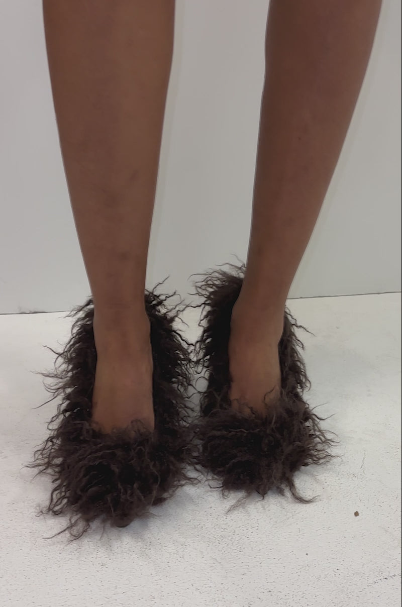 A video of a person wearing a dark brown leather skirt or coat and AZALEA WANG JAMEEKA CHOCOLATE POODLE HAIR PUMPS with stiletto heels stands against a plain white background, visible only from the waist down.
