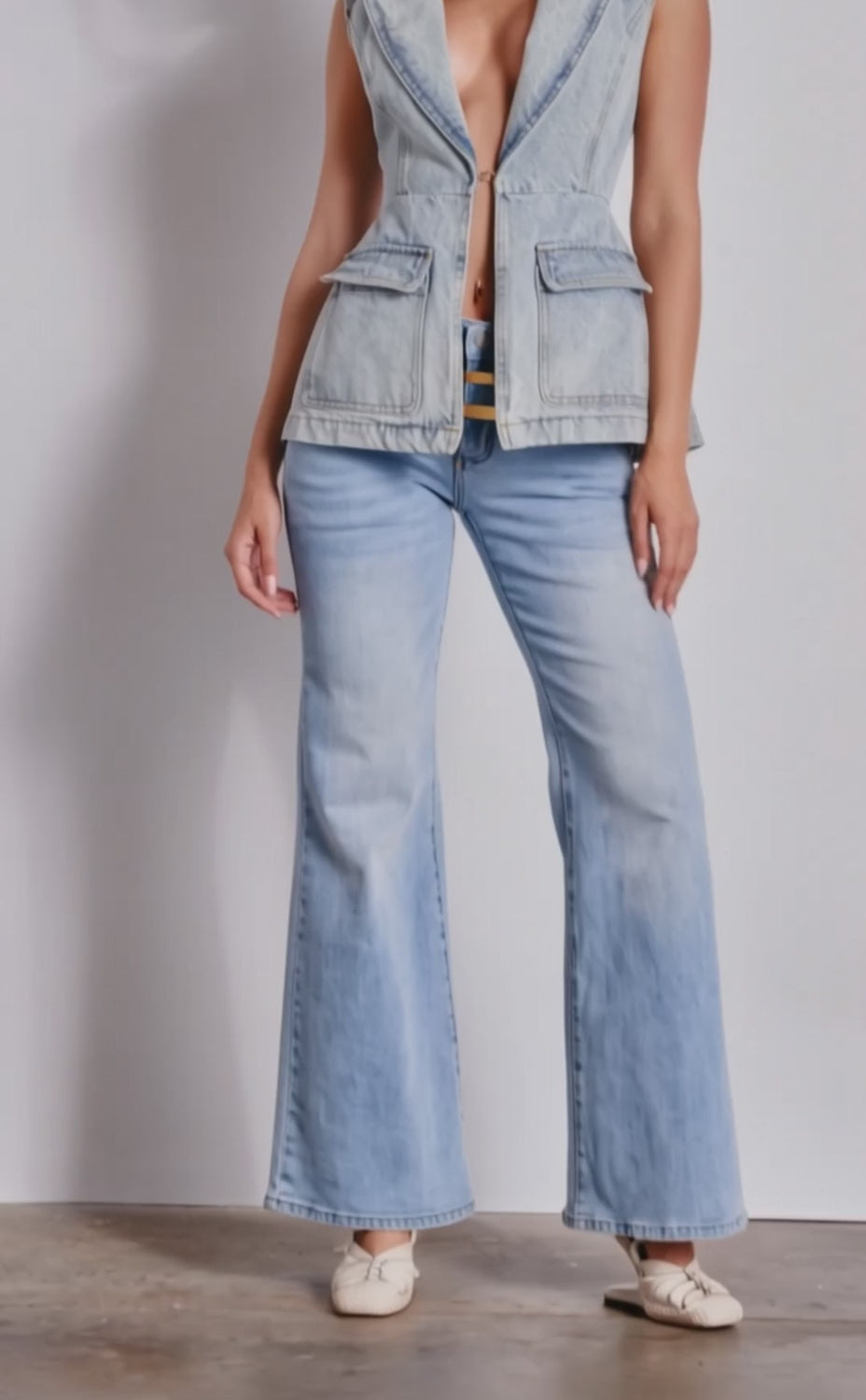 A video of a woman wearing the SCOUT DENIM BLAZER VEST, styled sleeveless with a deep V-neck, paired with wide-leg light wash denim jeans, white shoes, and statement earrings. 