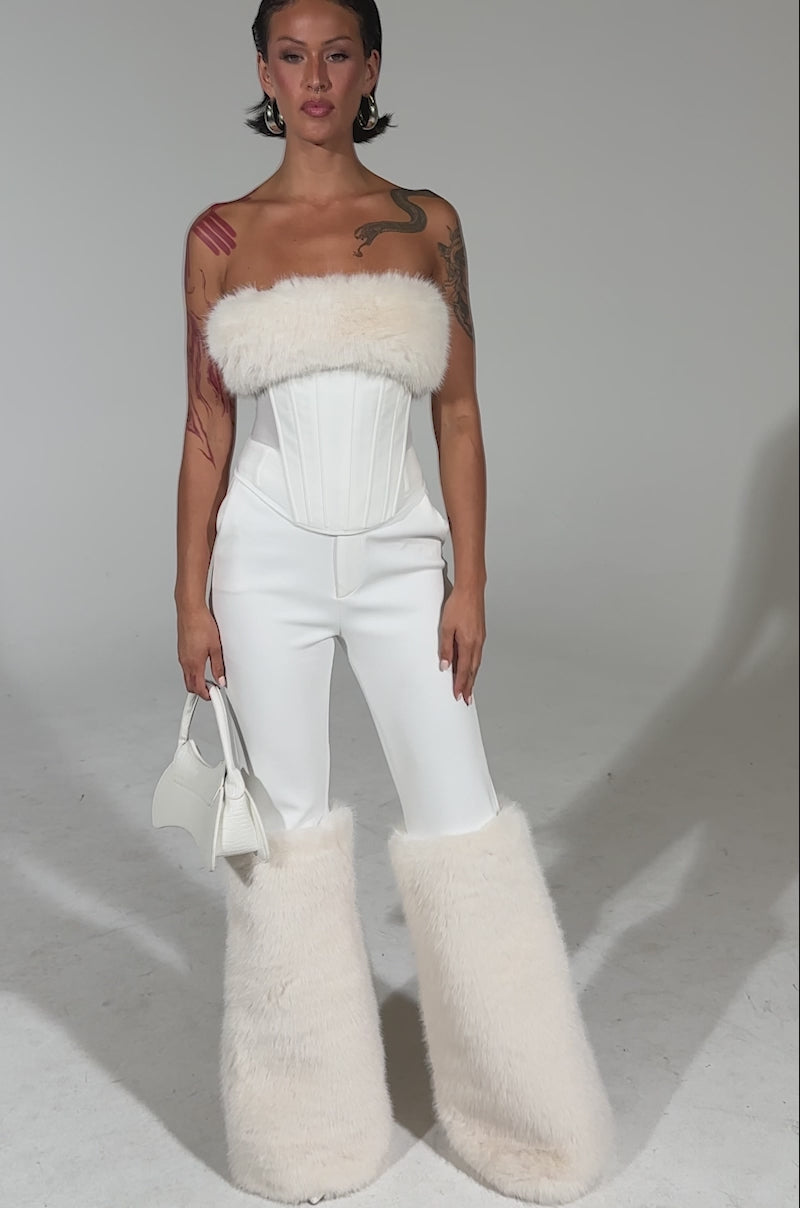 A video of an all-white look featuring the SNOW BUNNY CORSET WITH FUR by AKIRA Label, high-waisted pants, oversized fluffy leg warmers, and a small white handbag against a plain light background.