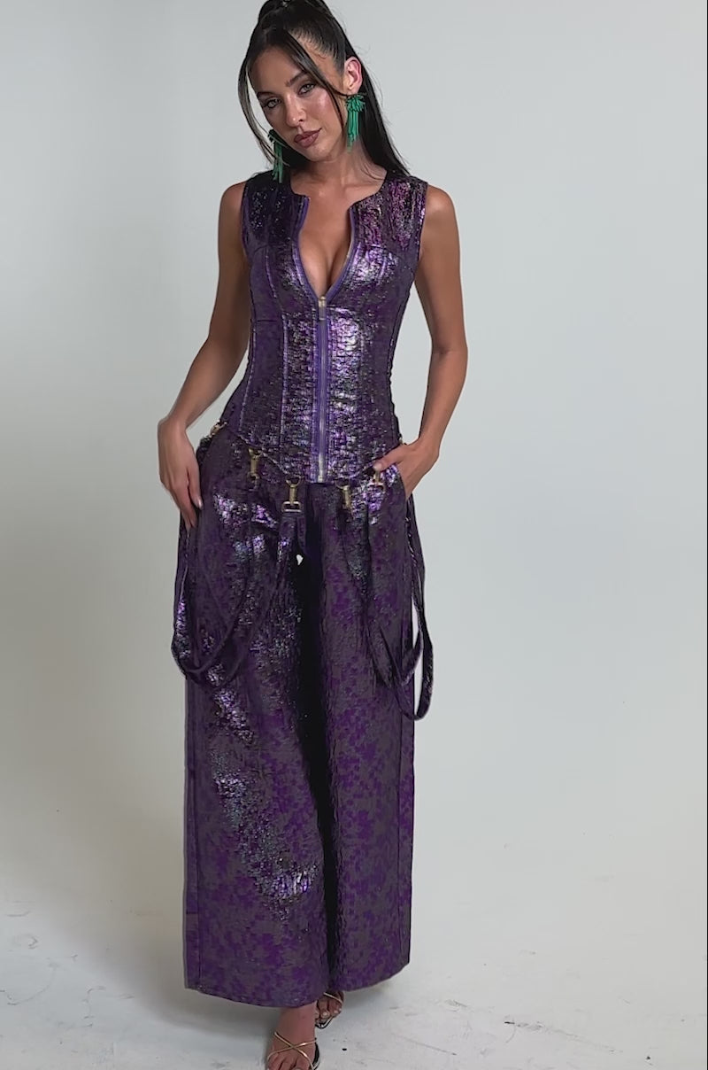 A video of a woman in the ROYALTY BROCADE CORSET TOP, a shiny purple textured sleeveless piece with a zip-up front and corset detail, styled with turquoise statement earrings and metallic high heels.