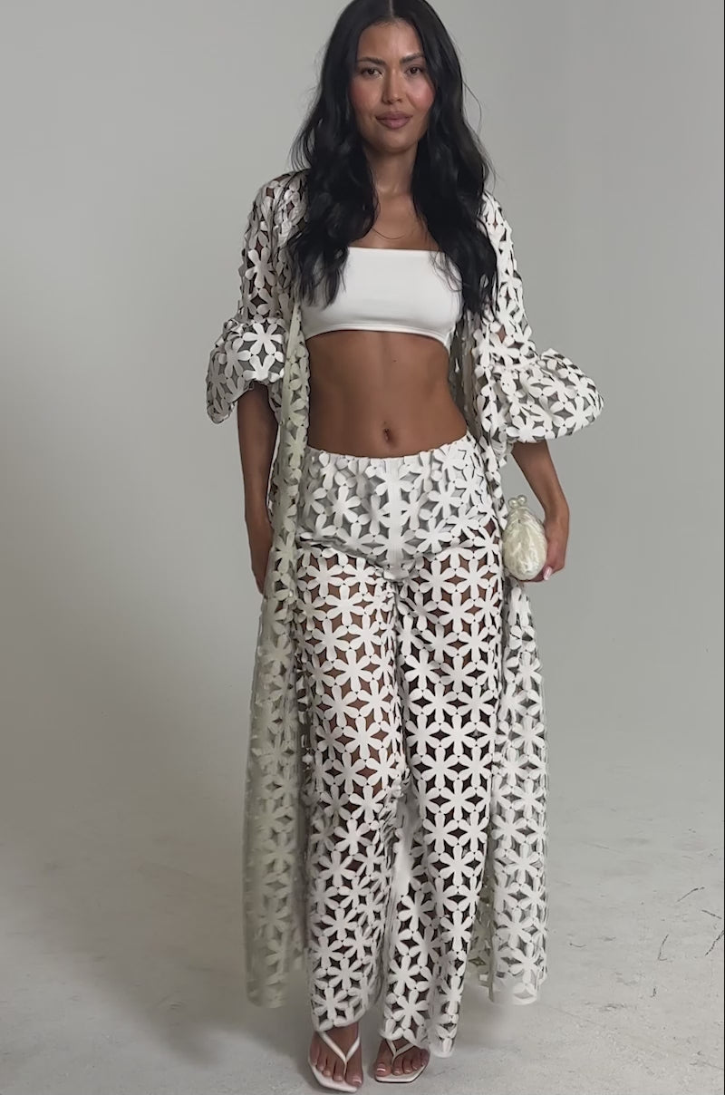 Video of woman modeling the LEAH TROUSER—high-waisted wide-leg pants with a geometric cut-out pattern. She accessorizes with a small clutch and heeled sandals for a chic look.
