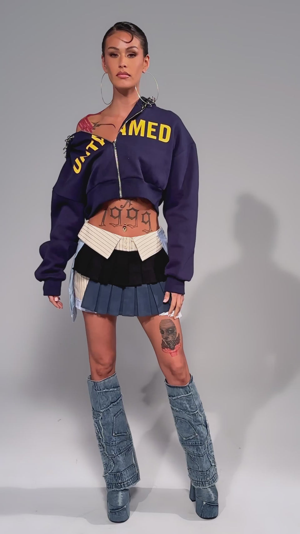 Video of woman modeling the STANDING ON BUSINESS MINI SKIRT, paired with a cropped blue menswear jacket with yellow lettering, tall denim boots, a small furry bag, and large hoop earrings that highlight their tattooed midriff.