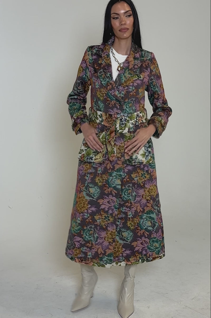 Video of woman modeling the SHOW OFF TAPESTRY TRENCH, a longline floral coat with a belt and pink, green, orange, and yellow flowers. She pairs it with light boots and a necklace