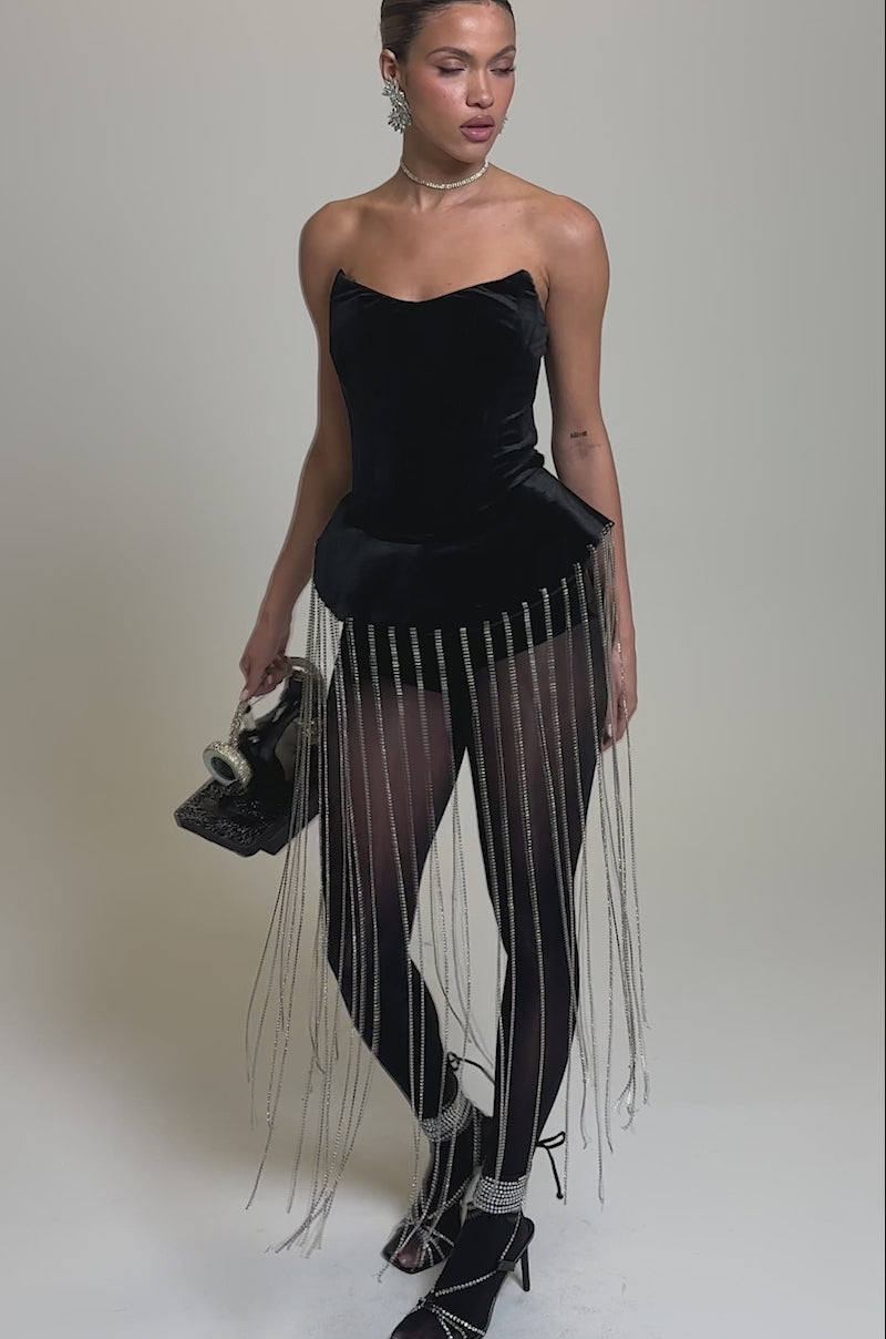 Video of woman modeling the PARTY 4 U VELVET FRINGE ROMPER—a strapless black piece with peplum and long silver chains—styled with black tights, heels, a box handbag, swept-back hair, and statement earrings for a chic disco look