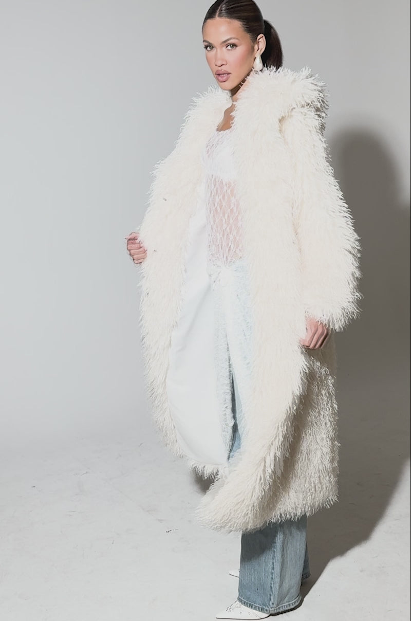 Video of woman modeling the SCARLETT FAUX FUR COAT over a sheer white lace top and skirt, styled with light blue wide-leg jeans and pointed-toe shoes, against a plain background.