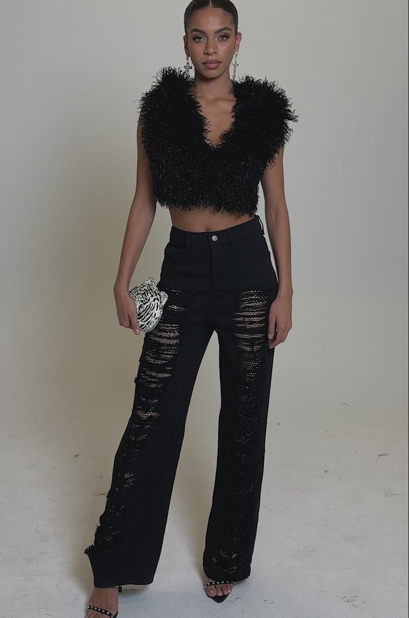 A video of a woman in CLARA DISTRESSED DENIM high-rise jeans with sheer, ripped detailing, a sleeveless black textured crop top, black heeled sandals, and holding a small silver clutch.