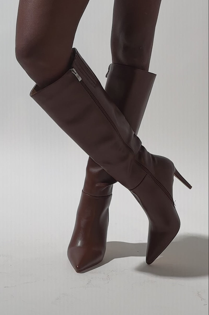 A video of a person wearing the AZALEA WANG HAILEYANN BROWN STILETO BOOT—knee-high, pointed toe, side zipper, brown faux leather with sleek fitted styling and elegant heels—paired with a blue skirt.