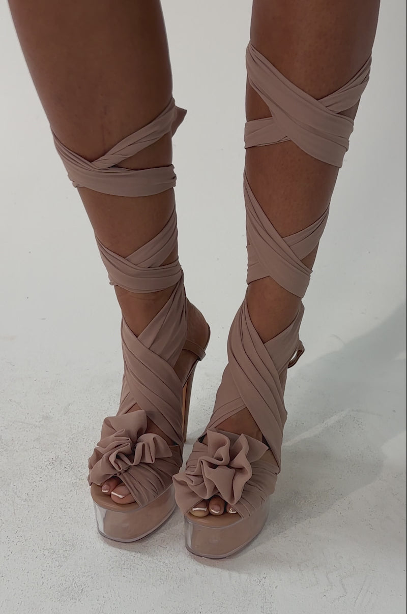 Video of woman modeling the AKIRA GOLD BEAUDEN TAN PLATFORM TIE SANDAL with square toe heel detailing, ribbons wrapped around crossed legs, against a plain light background.