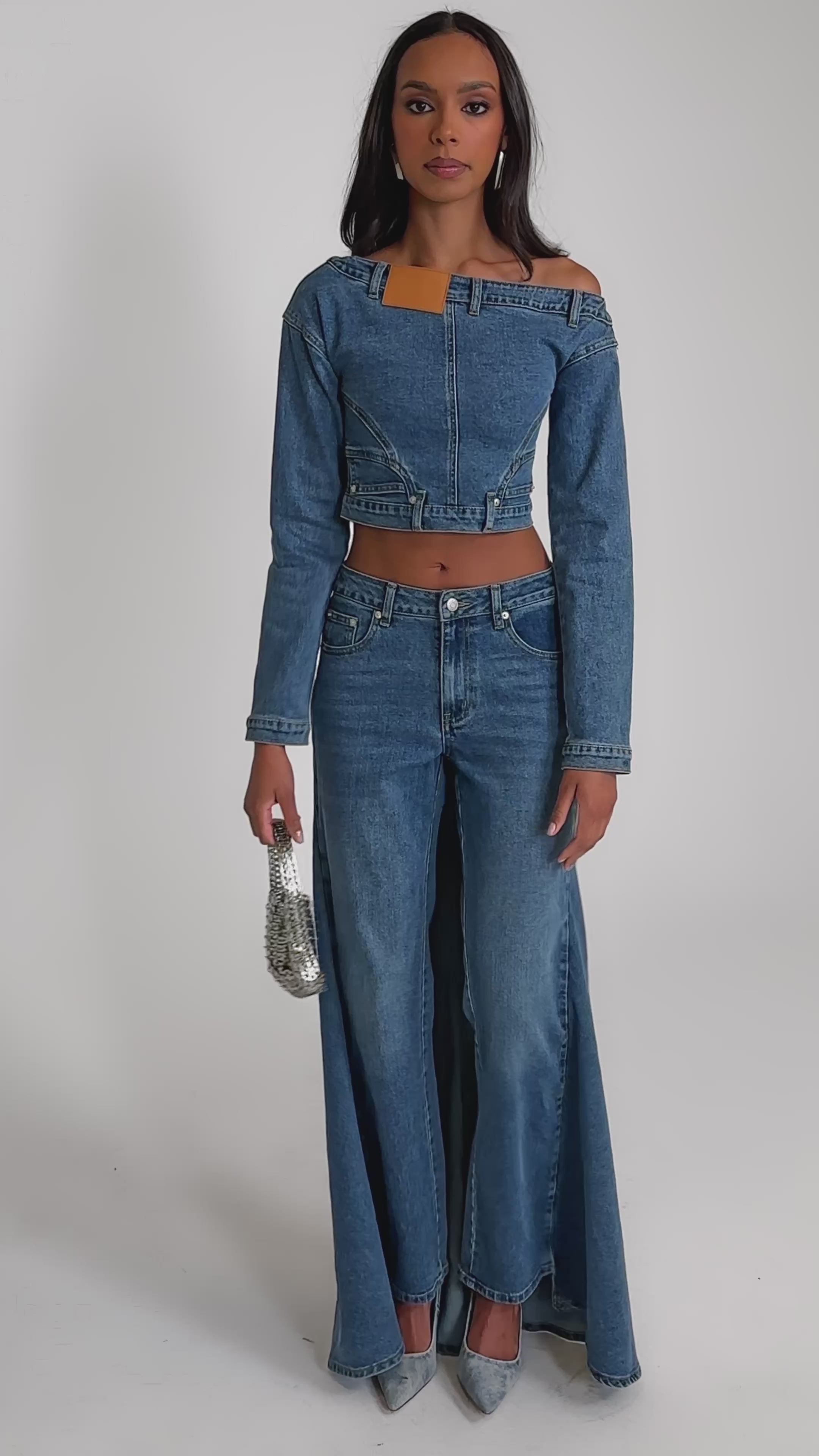 Video of woman modeling the SHAKE IT OFF DENIM OFF THE SHOULDER TOP with long sleeves and matching high-waisted denim pants, pairing it with a silver chain handbag for chic night out streetwear against a neutral backdrop.
