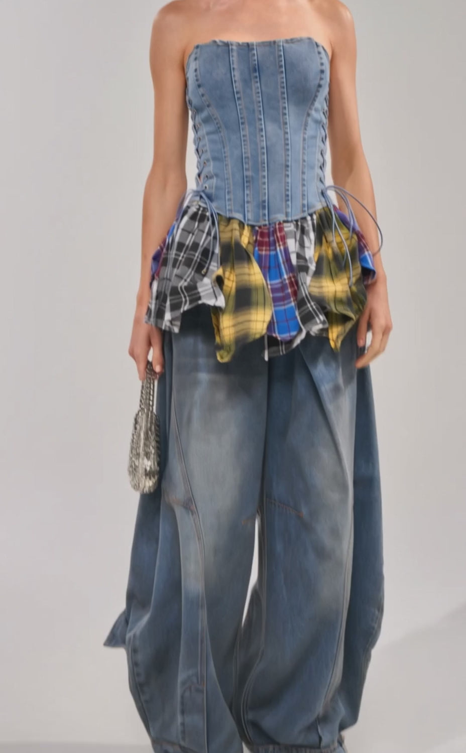 Video of woman modeling the MESS WITH ME PLAID DENIM CORSET, a strapless denim top with lace-up sides and a plaid hem, styled with wide-leg jeans against a plain light backdrop.
