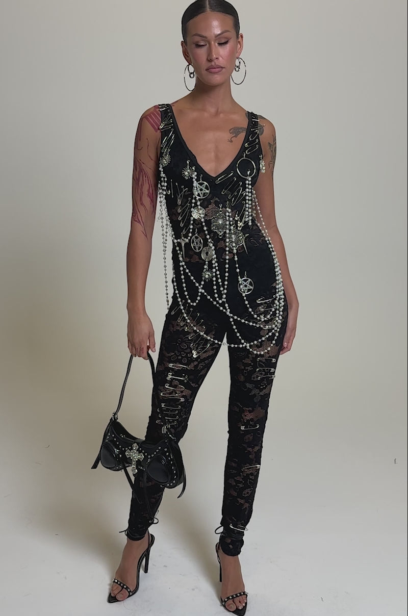A video of a person wearing the BABY I AM THE PARTY LACE JUMPSUIT—a curve-hugging, sheer floral lace piece adorned with chains and pearls—paired with black high-heeled sandals, large hoop earrings, and a small black handbag.