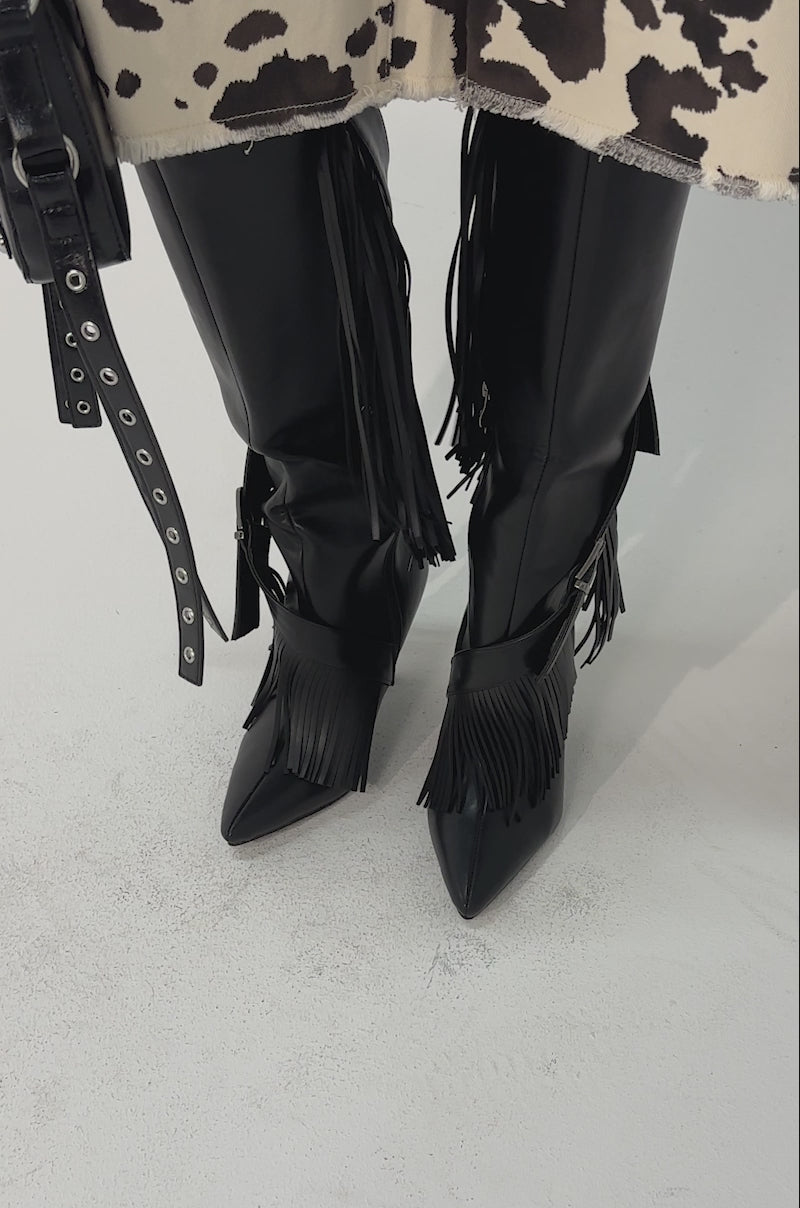 Video of woman modeling the AZALEA WANG DARELLE BLACK FRINGE BOOT with pointed toes, buckles and fringe sits 