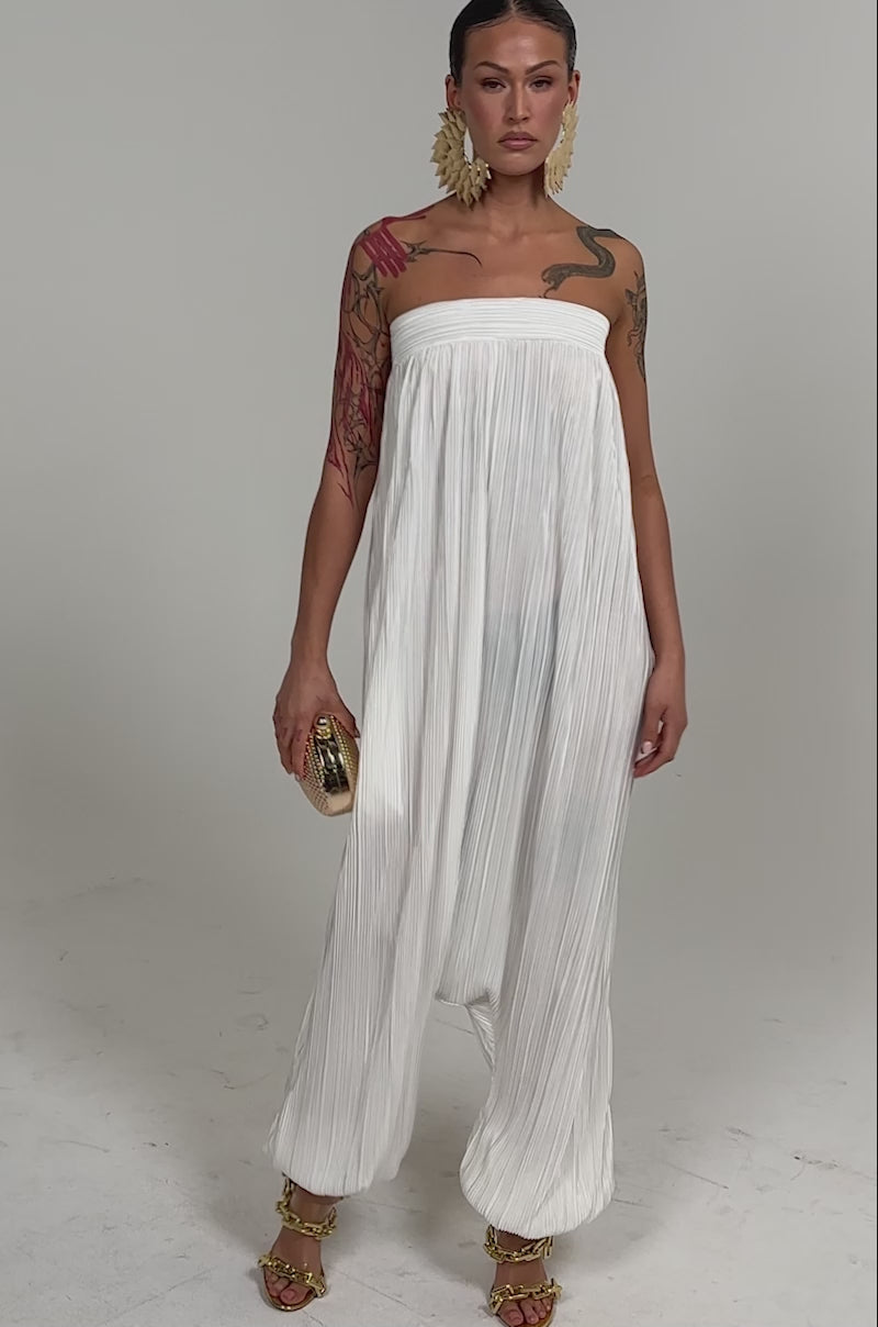Video of woman modeling the FROM THE CITY DROP CROTCH JUMPSUIT IN WHITE with flowing pleats, paired with gold sandals, large gold earrings, a textured clutch, and visible arm tattoos. Her hair is pulled back as she stands against a plain background.