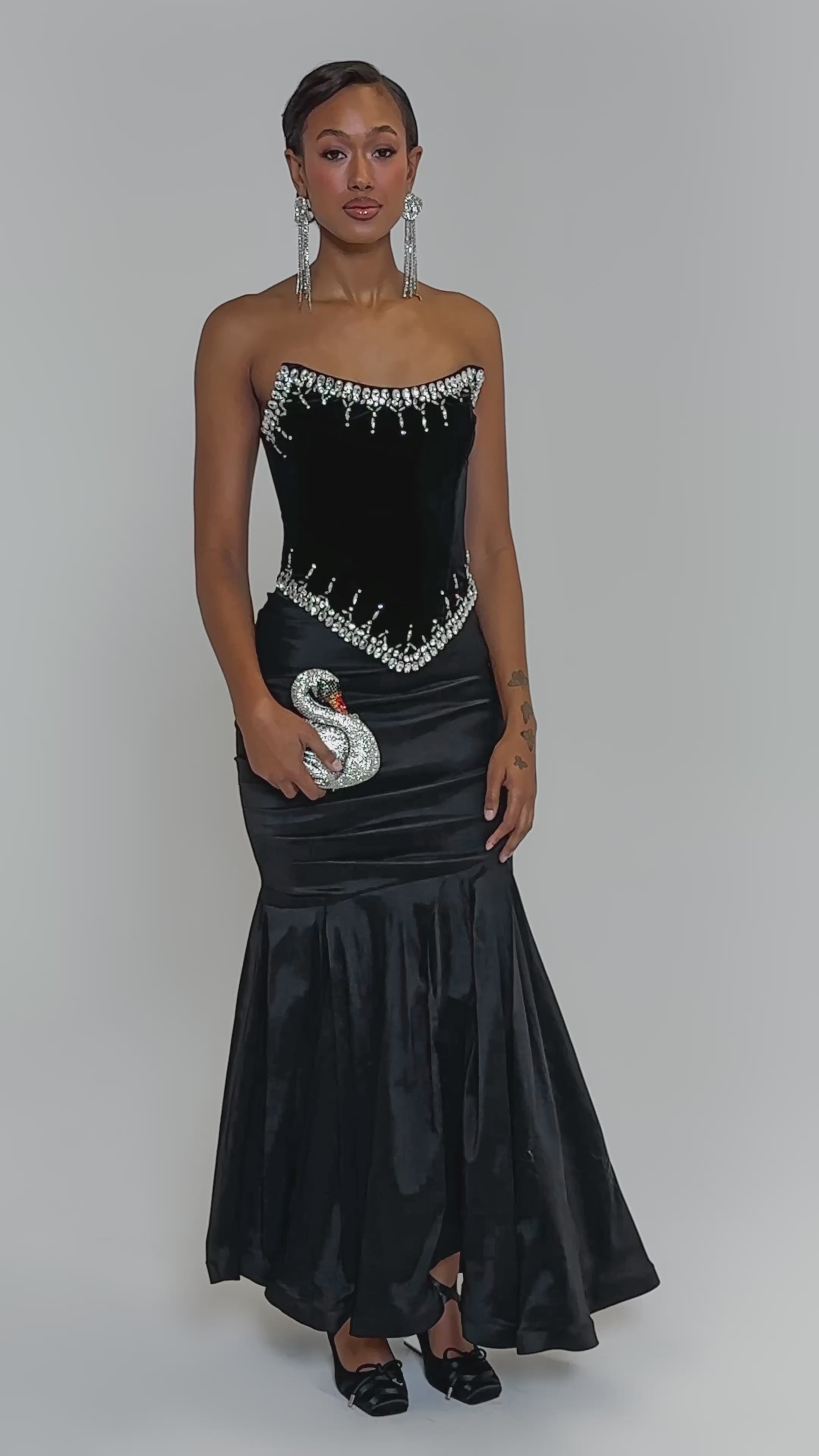 Video of woman modeling the LUX LIFE CORSET IN BLACK, a strapless velvet top with rhinestone accents at the neckline and waist. She has long dangling earrings and holds a glittery high heel, set against a plain light background.