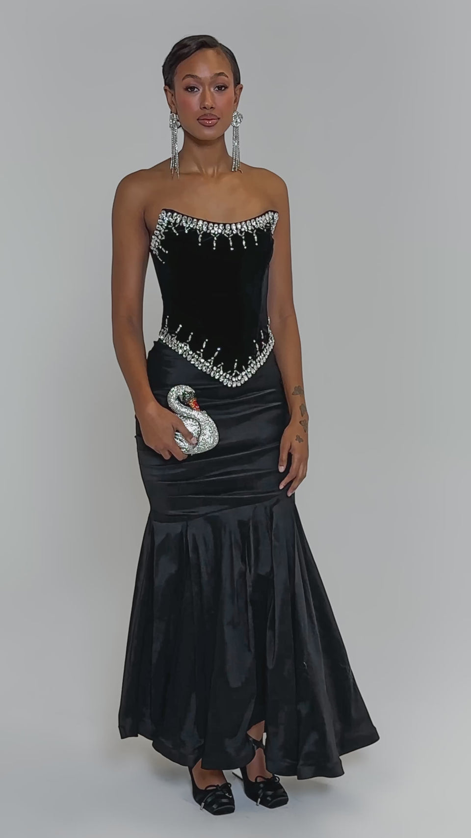 Video of woman modeling the LUX LIFE CORSET IN BLACK, a strapless velvet top with rhinestone accents at the neckline and waist. She has long dangling earrings and holds a glittery high heel, set against a plain light background.