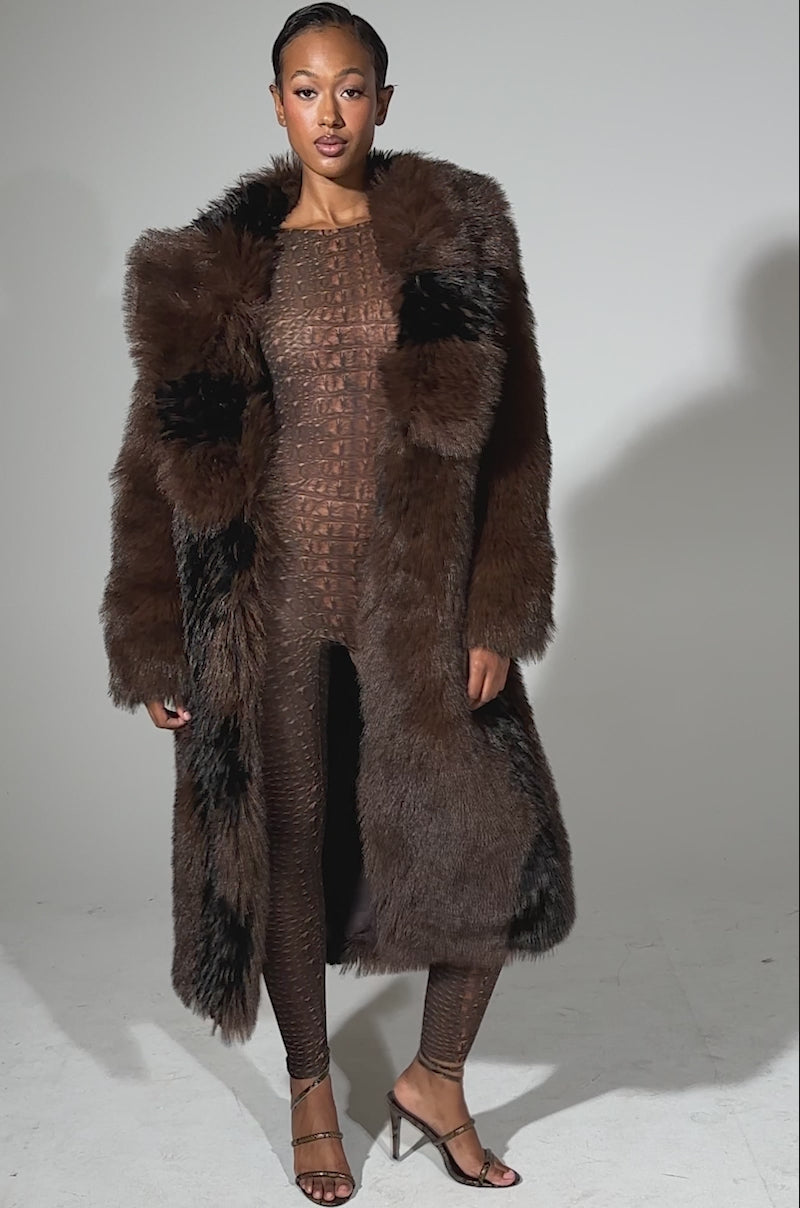 A video of a woman in dark sunglasses modeling the TELLURIDE FAUX FUR IN BROWN longline jacket over a fitted brown patterned jumpsuit and black strappy heels, set against a plain light background.