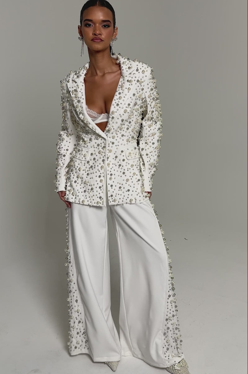 Video of woman modeling the KEEP IT GOING EMBELLISHED BLAZER with beaded pearls, paired with a white lace bra, white pants, and long earrings, set against a plain light background.