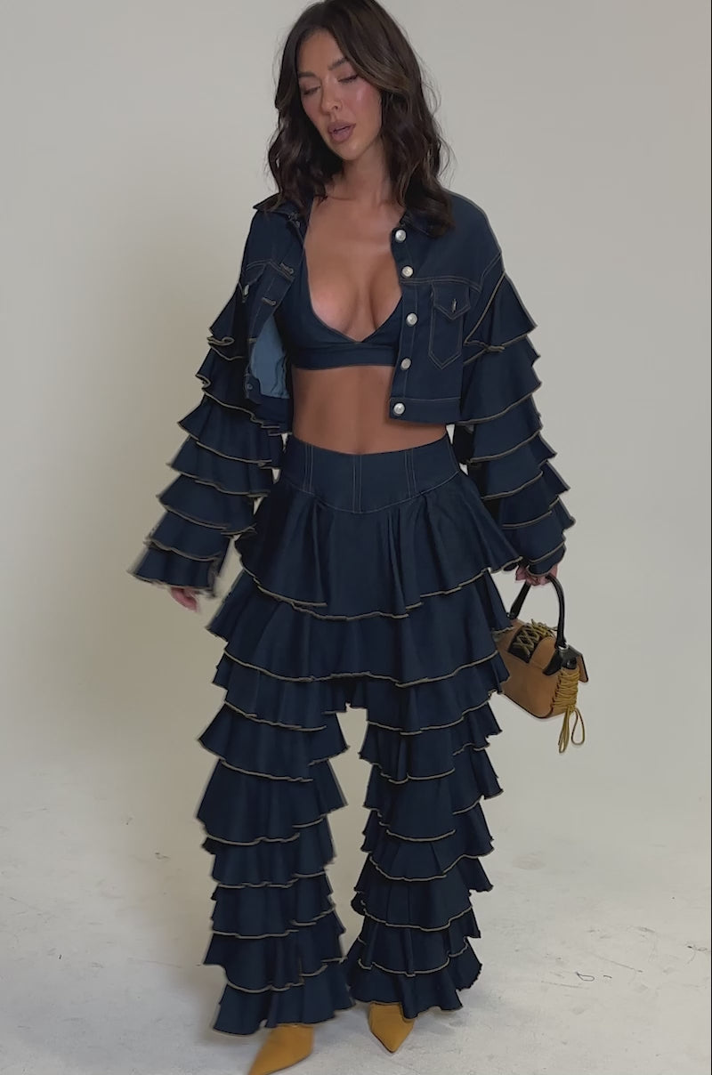 Video of woman modeling the SHAKE IT OUT CROPPED DENIM JACKET with tiered sleeves and a ruffled pant, she stands against a plain background, holding a small yellow handbag—creating an effortless statement layered look.