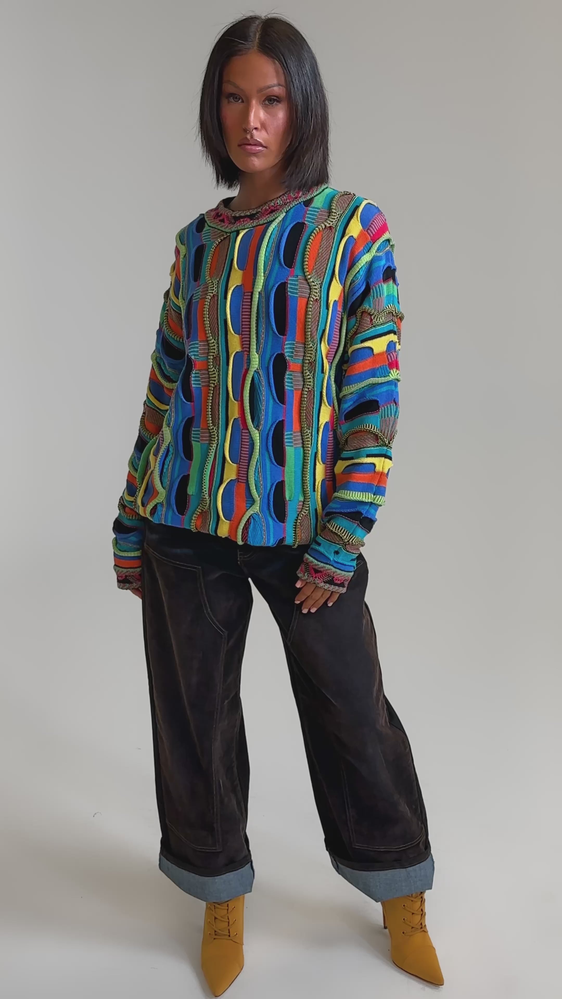 A video of a woman in the VINTAGE COOL OVERSIZED MULTI COLOR SWEATER, paired with loose dark cuffed jeans and mustard yellow lace-up boots against a plain light background.