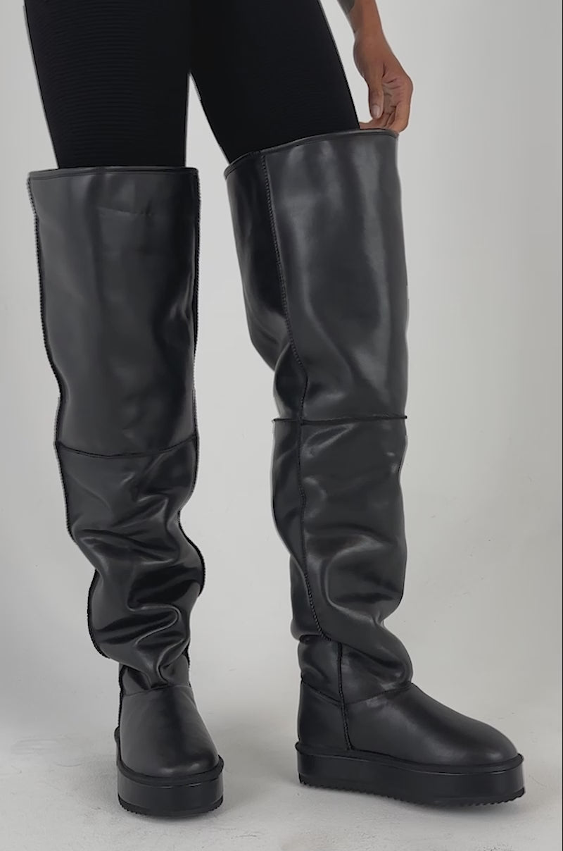 Video of a woman modeling the AZALEA WANG MARLOW BLACK PU FLATFORM OVER THE KNEE BOOT, lifting one leg to showcase the oversized black footwear.