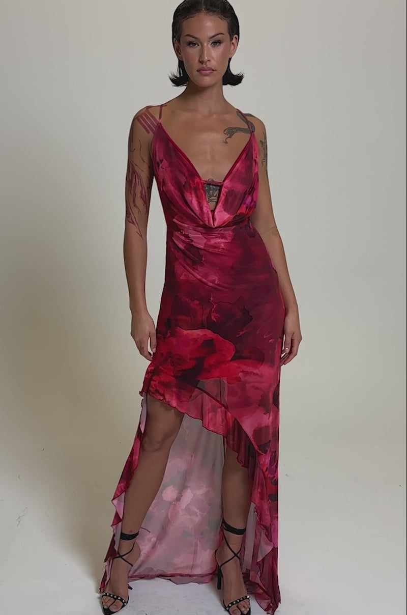 A video of a woman in the HILLARY FLORAL MAXI DRESS, a sleeveless asymmetrical maxi with a cowl neckline and sheer pink mesh, paired with strappy black heels. She has slicked-back hair and stands against a gray background.