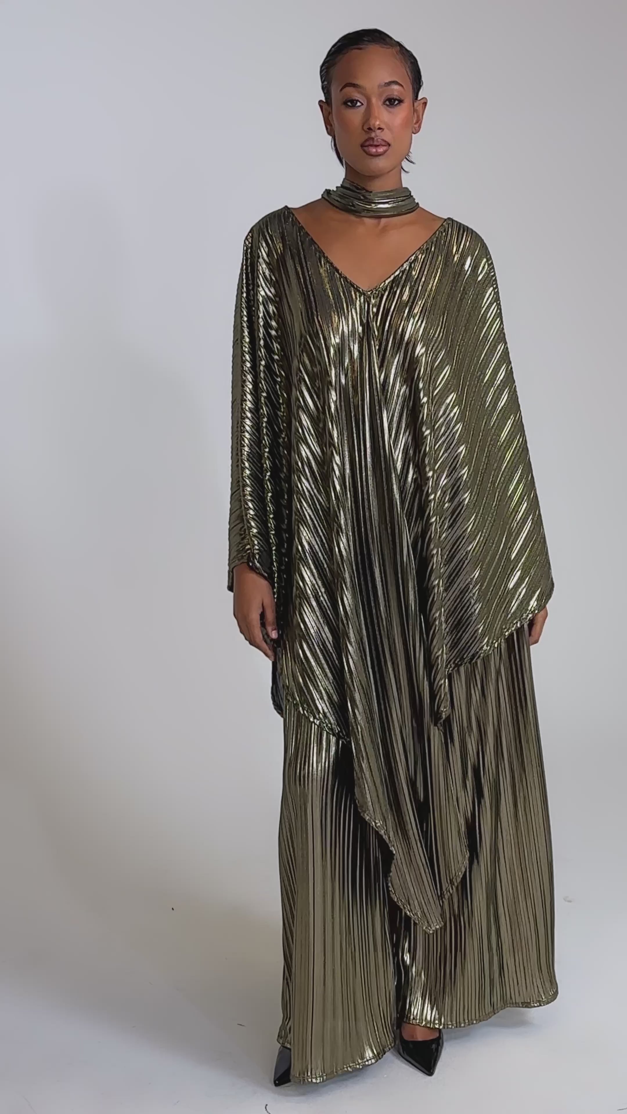 A video of a woman wearing the BEST DRESSED BLOUSE—a flowing, metallic pleated top with wide sleeves—paired with pants and black pointed heels, posing stylishly against a plain light background.