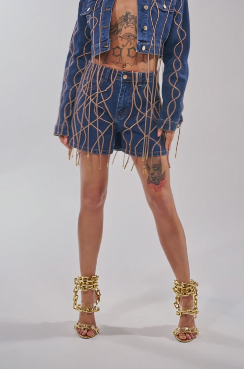 Video of woman modeling the NINA EMBELLISHED DENIM JACKET with matching shorts, both featuring gold chain details. She accessorizes with hoop earrings, layered necklaces, and displays chest and abdomen tattoos. Her dark hair is slicked back.
