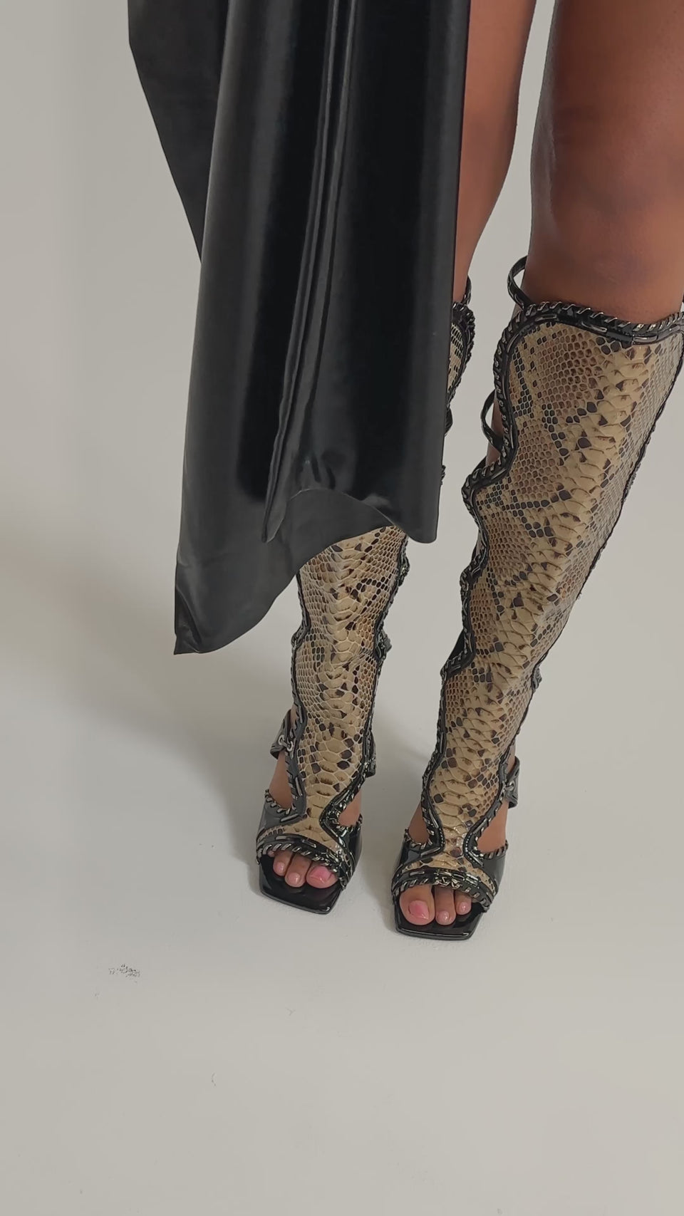 A video of a person in the AKIRA GOLD INDRANI BLACK HIGH SHAFT GLADIATOR SANDAL, featuring a bold knee-high silhouette with black and tan snakeskin pattern, wavy black trim, and an open-toe design against a light background.