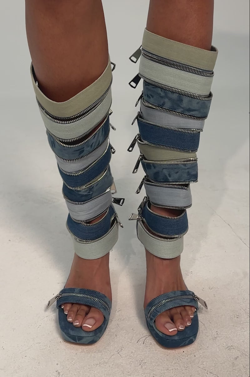 A video of a model wearing the AKIRA GOLD ONREPEAT DENIM EMBELLISHED PLATFORM STILETTO SANDAL BOOT. It is a knee-high, open-toe denim boot with multiple horizontal zippers and an ankle strap, finished with a sleek stiletto heel and statement platform.