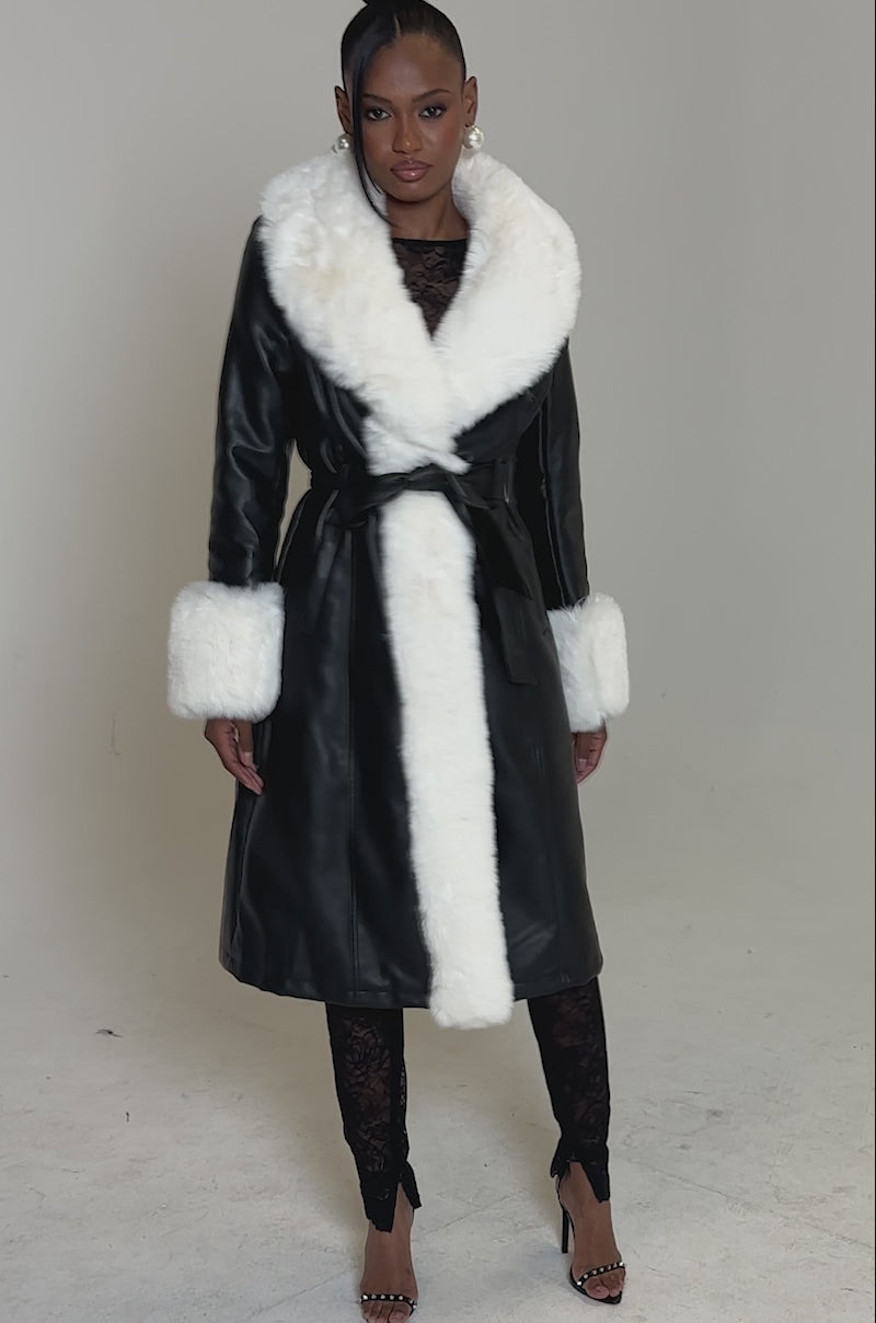 Video of woman modeling the SNOW BUNNY FUR TRIM TRENCH, a black belted coat with white fur collar and cuffs, paired with black lace pants and open-toe heels, set against a plain light background.