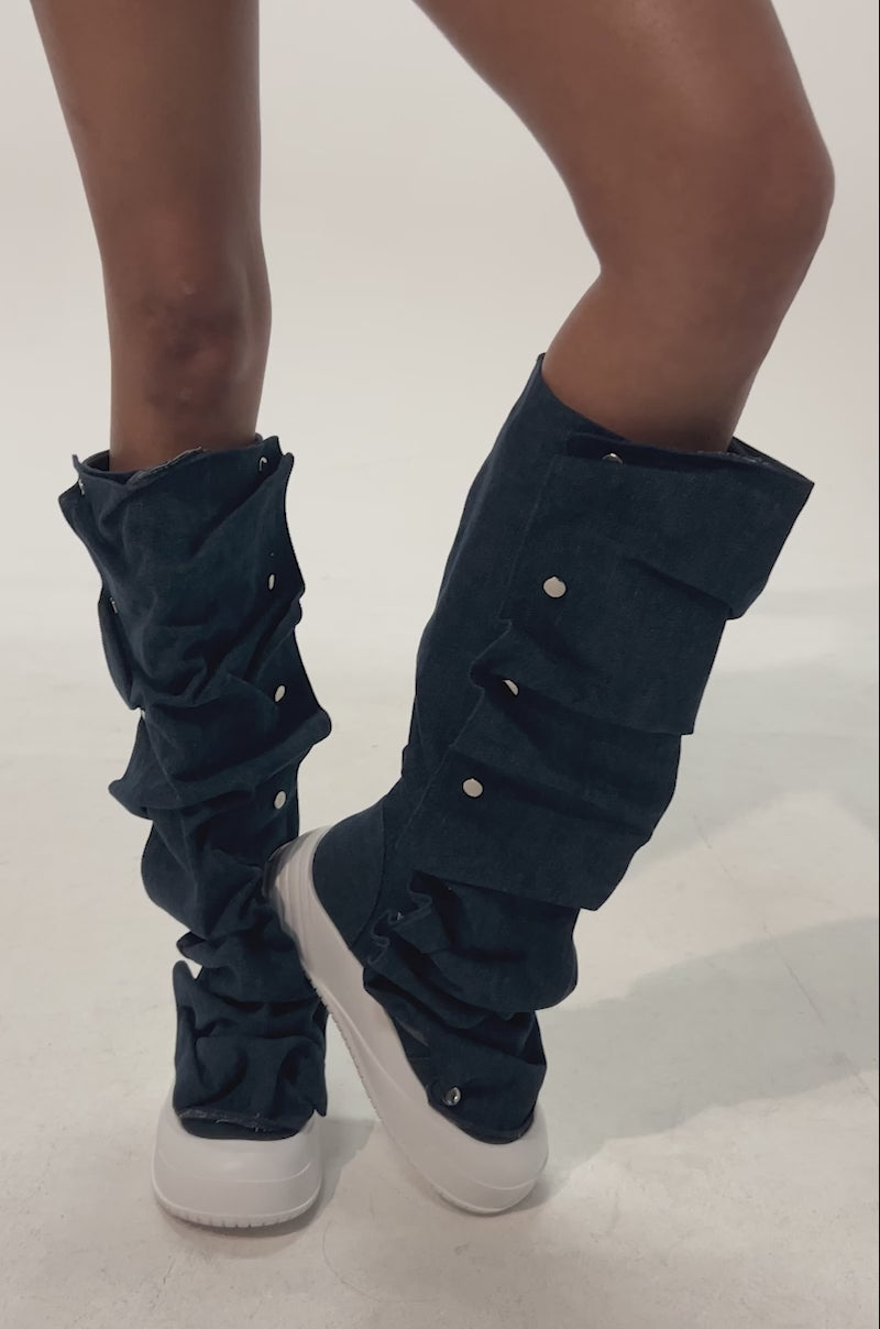 Video of woman modeling the AKIRA GOLD ALIXY DENIM FLAT RUFFLE BOOTS with frayed denim shorts and white sneakers, creating a casual streetwear vibe with the unique slouchy leg covers and white buttons against a plain background.