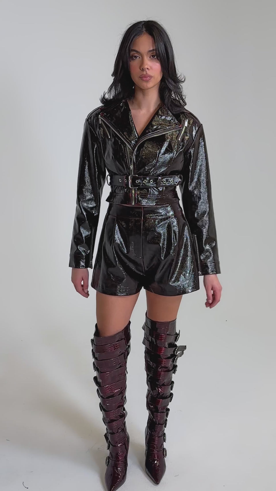 A video of a woman wearing the OH HOT DAMN FAUX LEATHER SHORT in shiny dark brown vegan leather, paired with a matching jacket and tall, textured knee-high boots. 