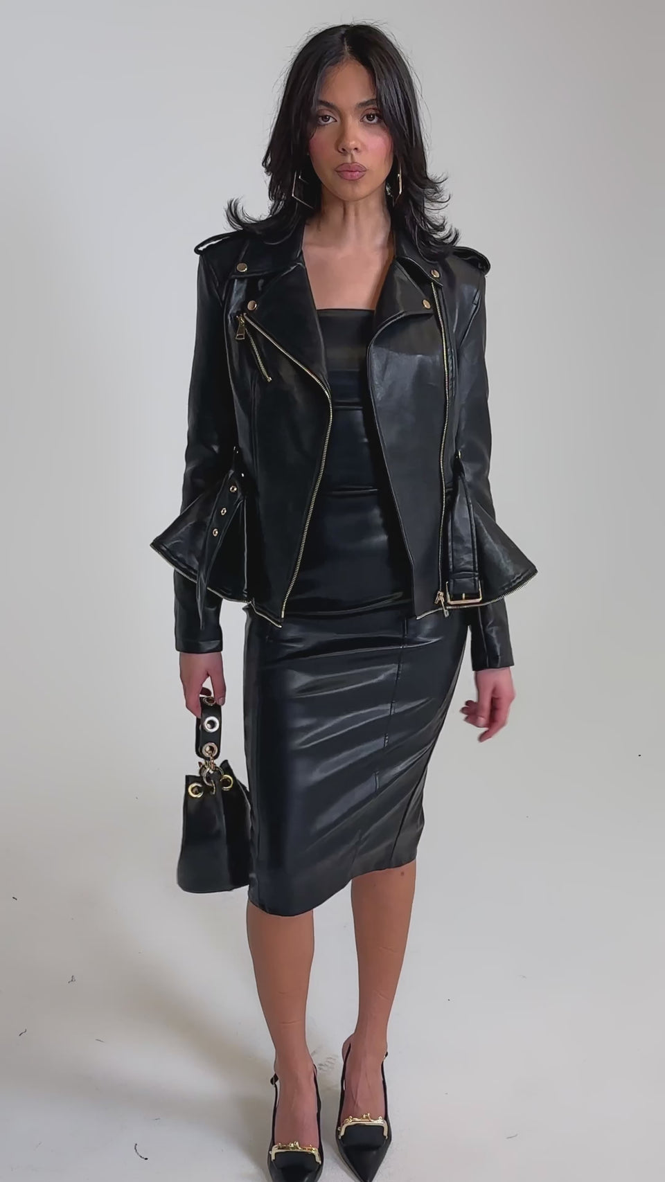 A video of a woman in the JULES PEPLUM MOTO jacket with an asymmetrical zip, paired with a black leather skirt, pointed gold-accented heels, and a small black handbag. 