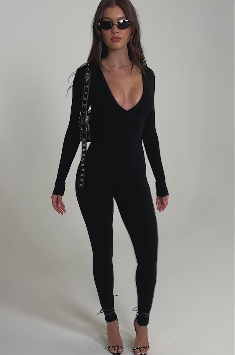 A video of a woman wearing the DEEP V LONG SLEEVE JUMPSUIT, a fitted black ribbed knit piece with a sculpted bodice and deep V-neck. 