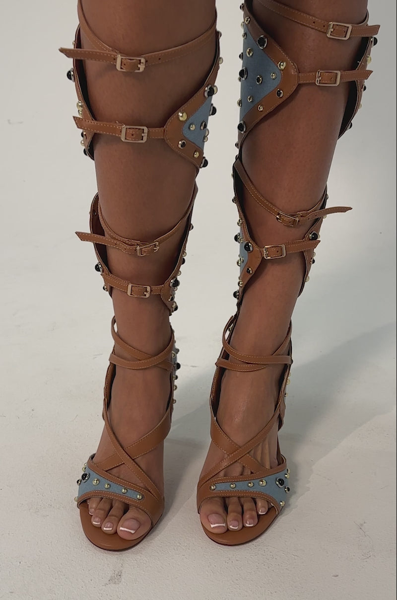 Video of woman modeling the AKIRA GOLD LULANA DENIM METAL STUDDED GLADIATOR STILETTO SANDALS with tan leather straps, blue panels, and gold studs.