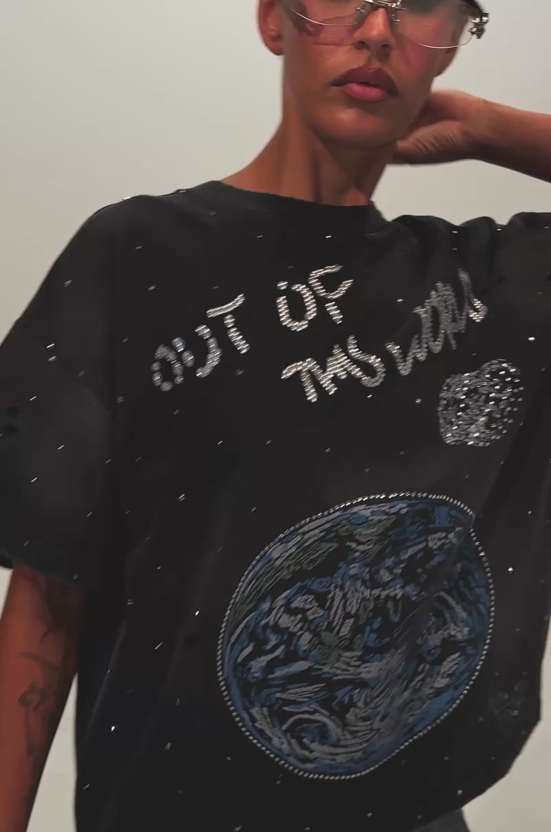 Video of a woman modeling the OUT OF THIS WORLD GRAPHIC TEE, which features a space-inspired design of Earth, the Moon, and the phrase "Out of My World," stands facing forward with one hand in her pocket.