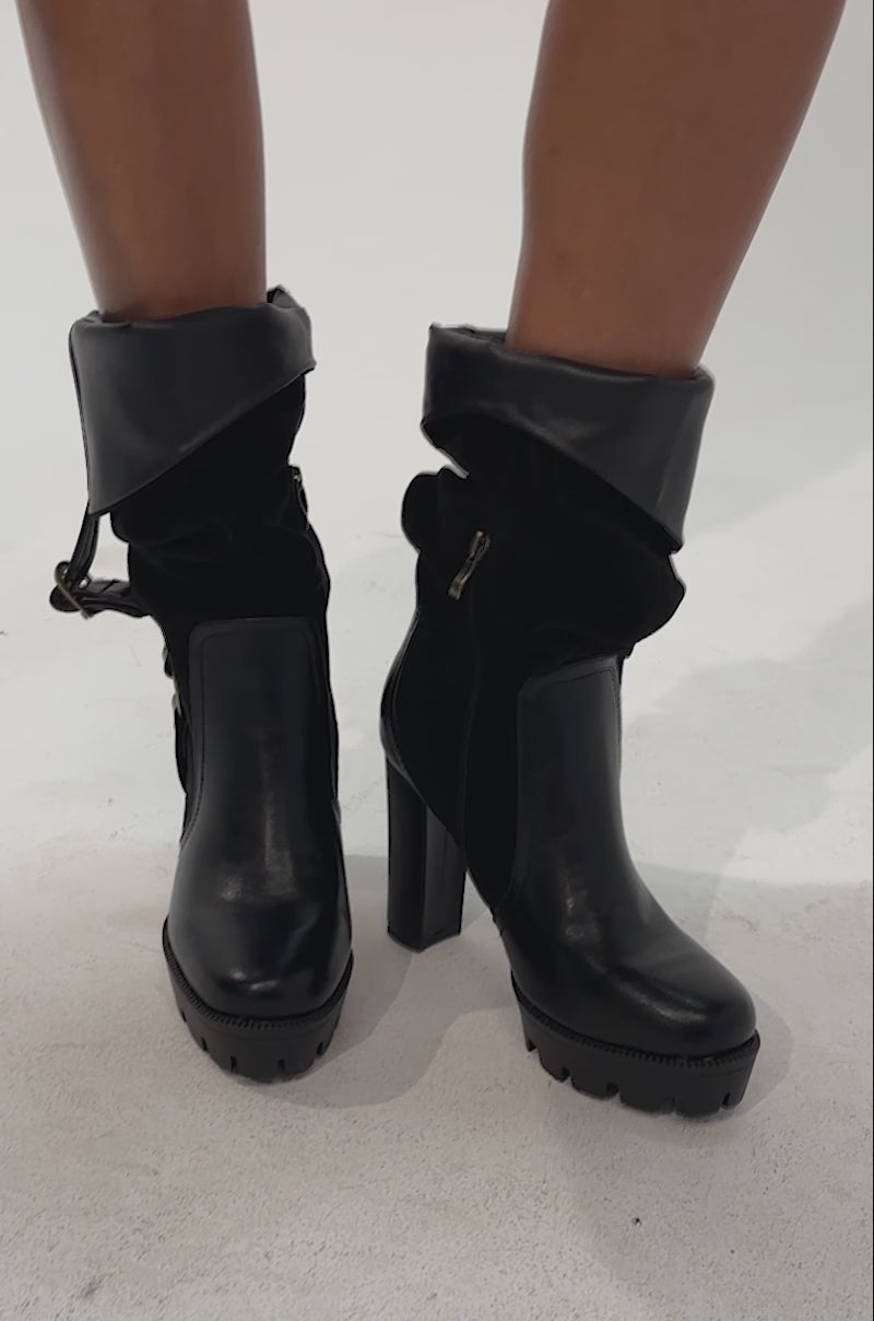 Video of a woman modeling the AZALEA WANG STREAMWOOD BLACK FAUX SUEDE BLOCK SLOUCHY BOOTIES with a modern silhouette, and holding a small black handbag stands against a plain light background.