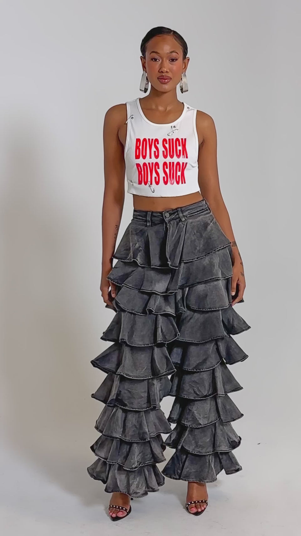 A video of a woman wearing the BOYS LIE Distressed Cropped Tank in White with bold red lettering and high-waisted gray pants, accessorized with large hoop earrings.