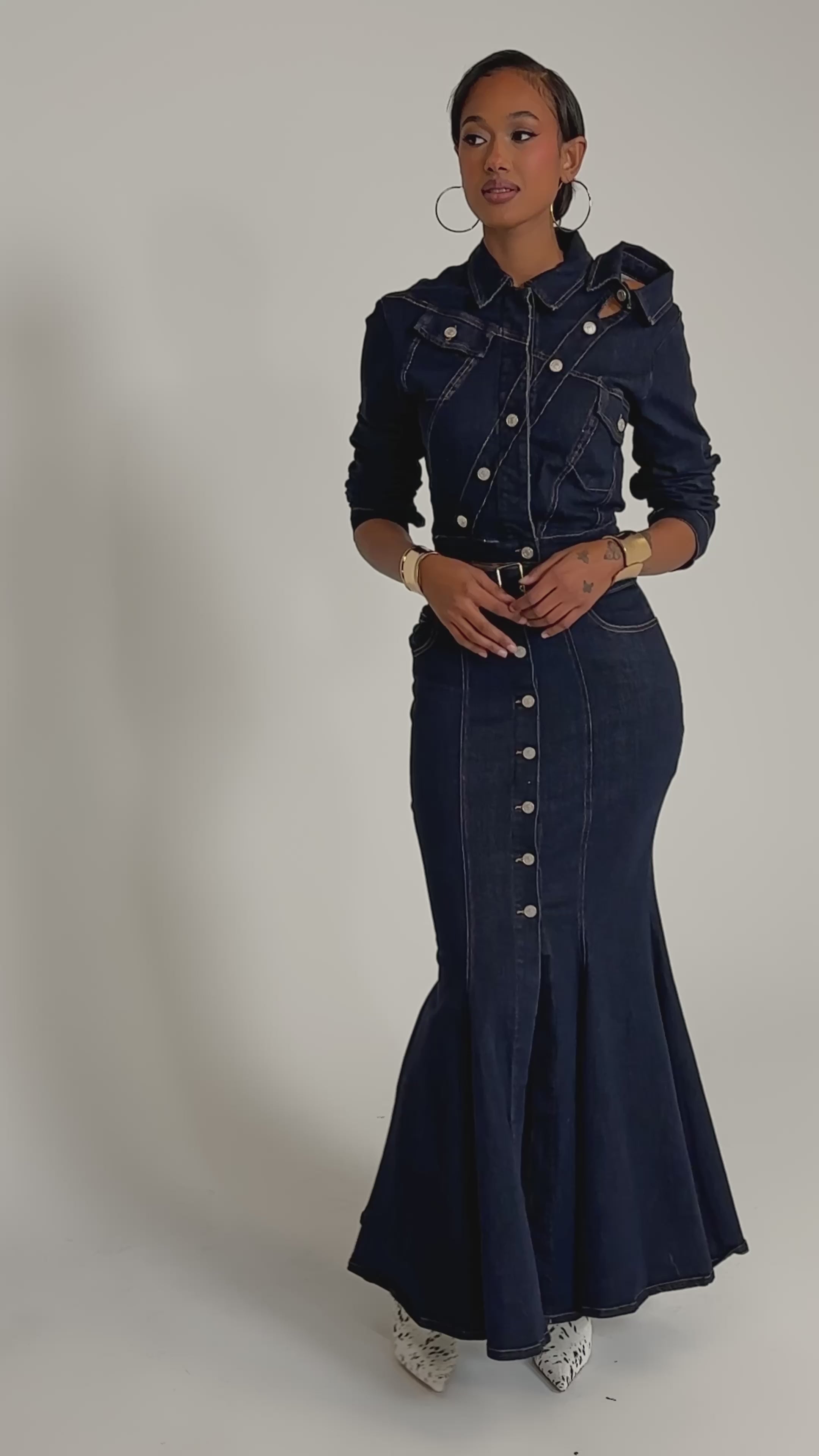 A video of a woman in the YOU GOT ME SPINNING DENIM BUTTON DOWN, an indigo wash fitted set, styled with hoop earrings, gold bracelets, and white patterned ankle boots against a plain background.