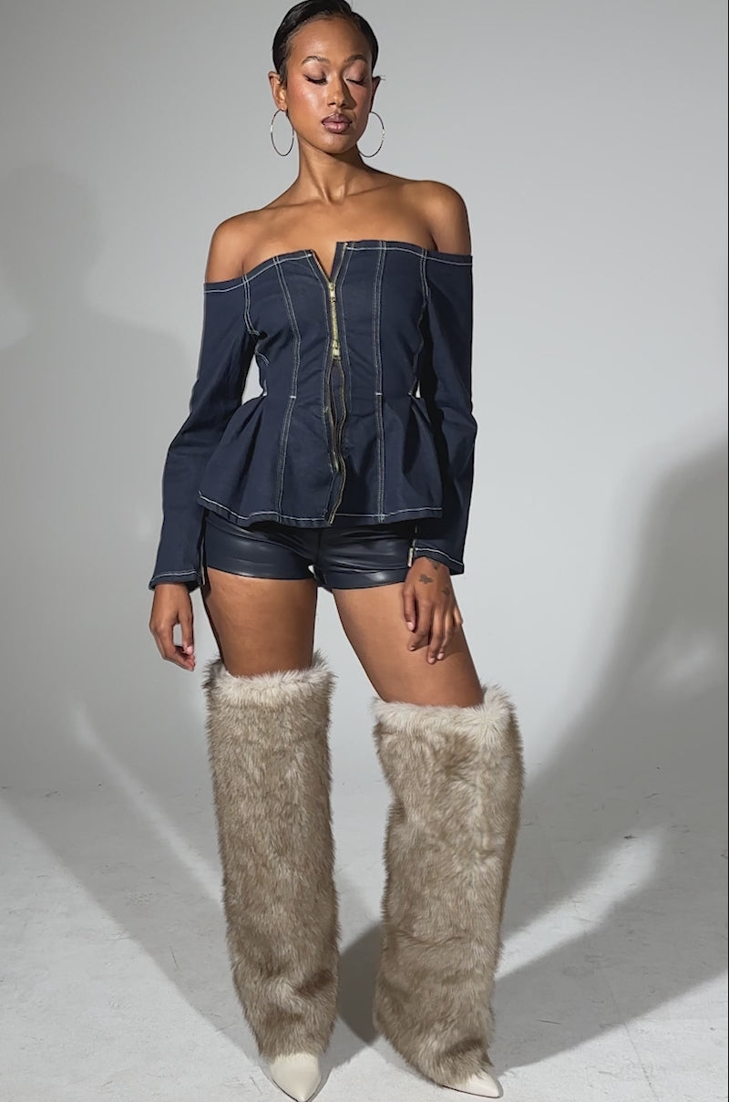 A video of a woman standing against a light background wearing the DENIM FOR DAYS ZIP UP BLOUSE, black shorts, and tall tan faux fur boots. She accessorizes with hoop earrings and has her hair styled back.