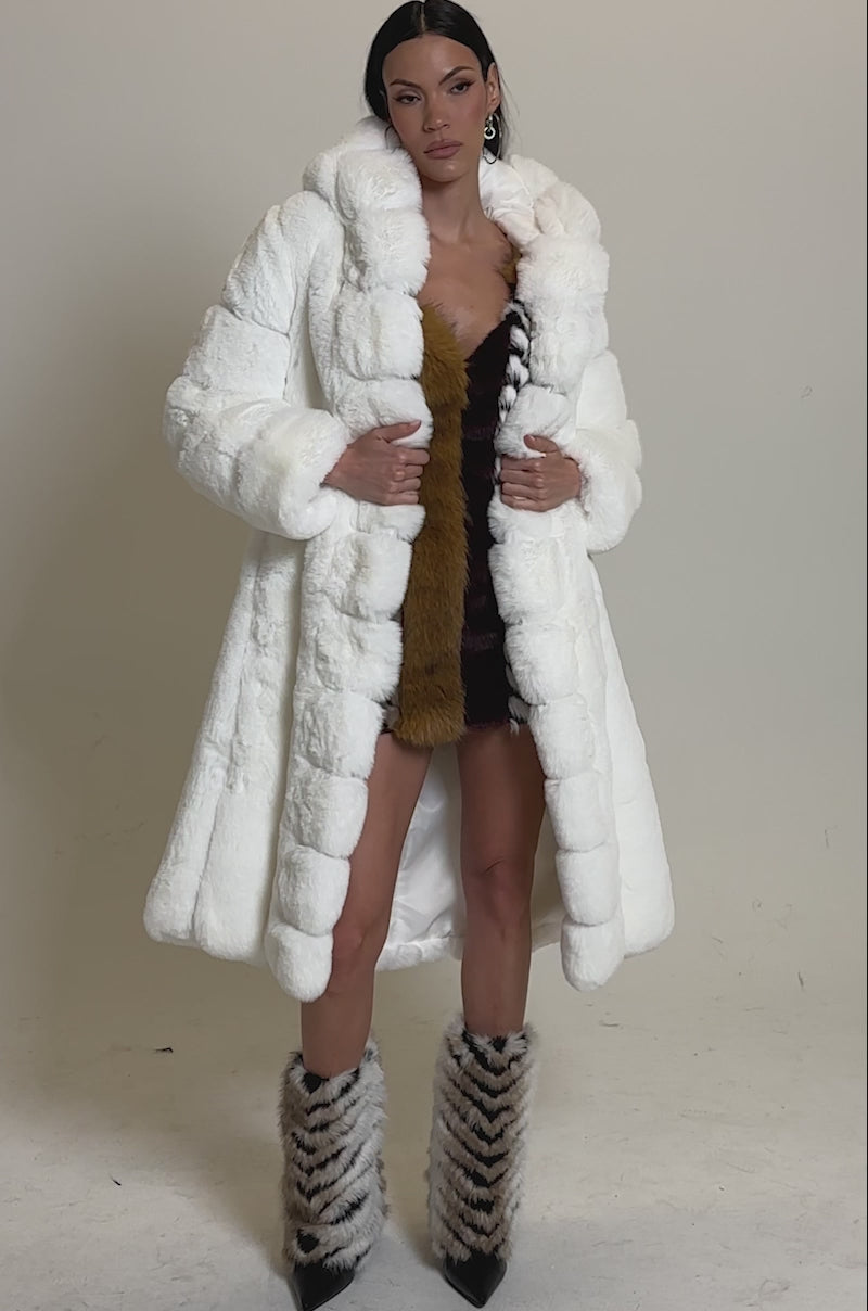 A video of a woman in the BERLIN HOODED FAUX FUR IN WHITE coat, draped over her shoulders, styled with a short multicolored faux fur dress and matching boots, highlighting stylish winter outerwear against a plain light background.