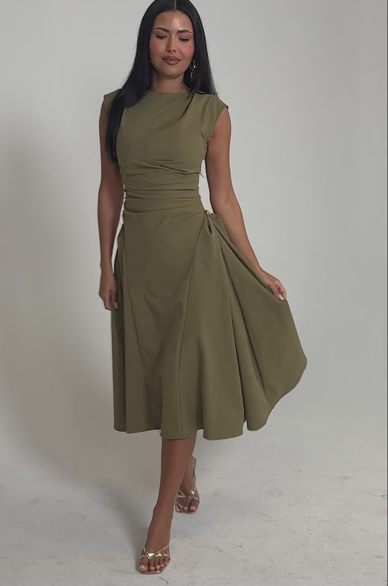 Video of woman modeling the IT'S MY TIME MIDI DRESS IN OLIVE, featuring a high neck and ruched details, paired with gold heels and a gray snakeskin clutch as she smiles at the camera against a light background.