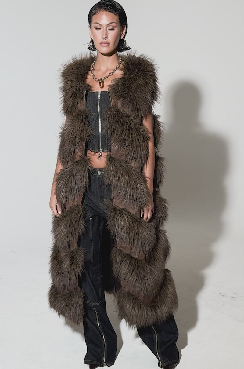 Video of woman modeling the CAN'T BE BOTHERED PANELED FUR VEST, a brown faux fur longline piece, layered over a dark cropped top and wide-leg zippered pants, set against a plain light background.