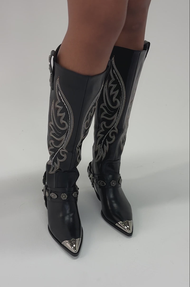 Video of woman modeling the AZALEA WANG GERVASE BLACK PU SILVER HARDWARE WESTERN BOOT with silver hardware and harness-style straps standing on a light-colored surface.