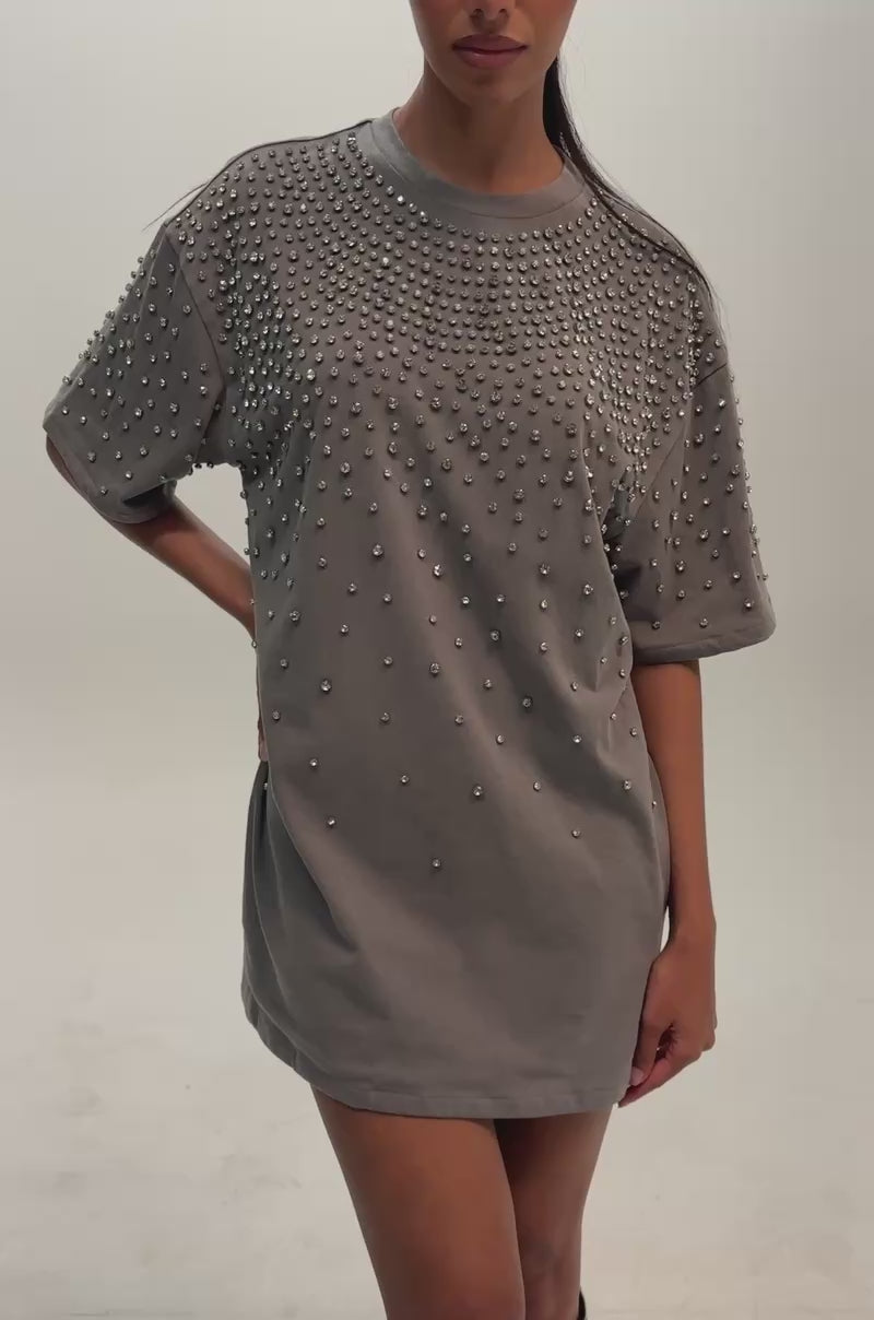 Video of a woman modeling the IN MY ELEMENT RHINESTONE TEE SHIRT MINI DRESS, a loose gray dress embellished with small metal studs across the shoulders and chest. 