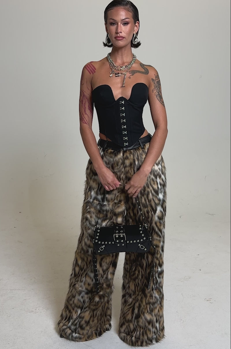 A video of a woman in a black strapless corset top and the CATCH ME IF YOU CAN LEOPARD FUR TROUSER, accessorized with a black studded handbag, statement jewelry, and slicked-back hair against a plain background.