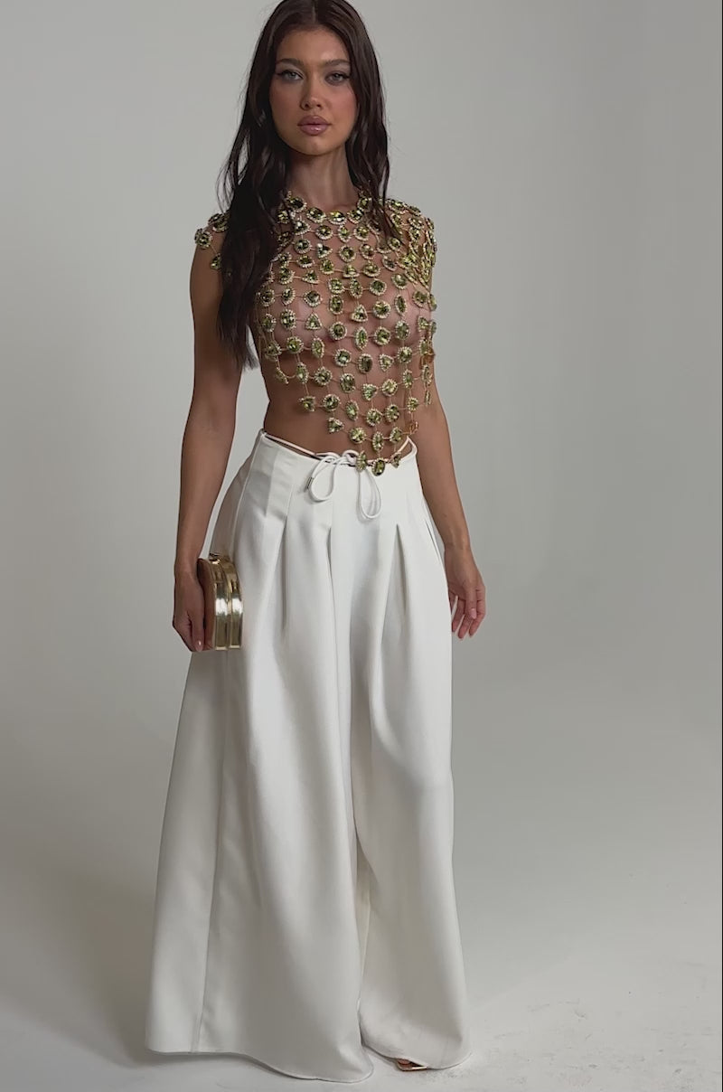 Video of woman modeling the ALL I NEED IS MONEY RHINESTONE CHAIN TOP over a nude underlayer, paired with high-waisted white pleated trousers, standing against a plain light background.