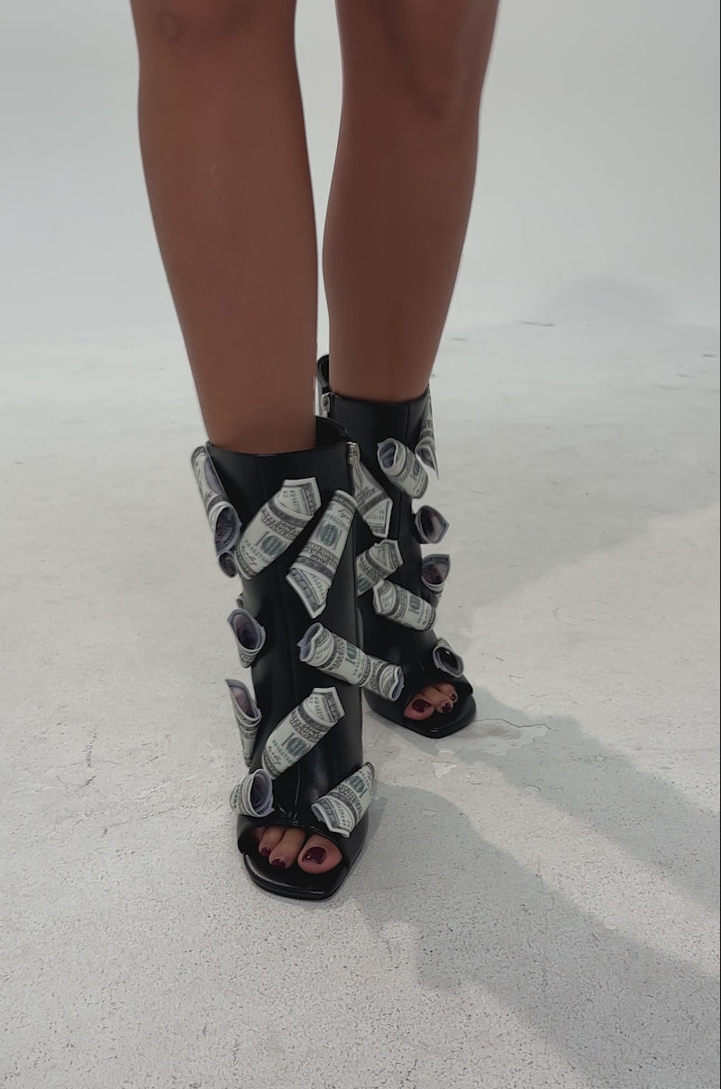 A video of a person wearing the AZALEA WANG DINERO BLACK MONEY OPEN TOE BOOTIE, featuring black stiletto heel sandals decorated with faux dollar bills, paired with a white skirt with black trim, standing against a white background.
